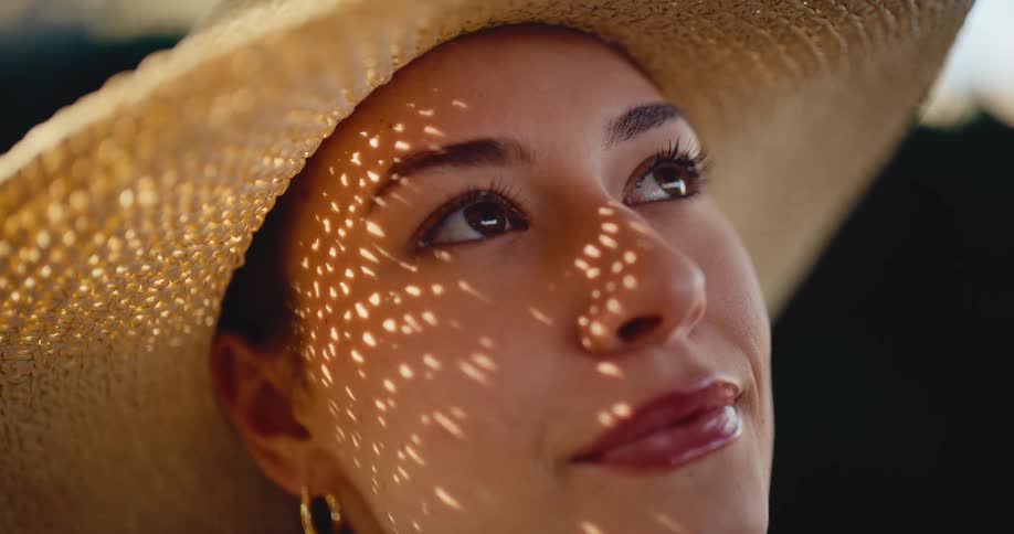 Sunlight filters through a woman's straw hat, creating a beautiful dappled shadow pattern on her serene face