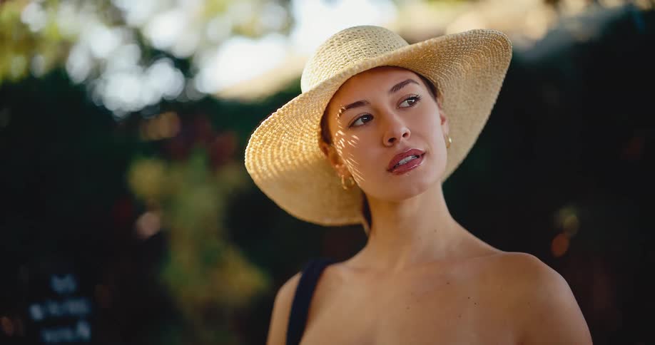 An elegant woman in a large straw hat stands thoughtfully in the soft, dappled sunlight of a lush garden