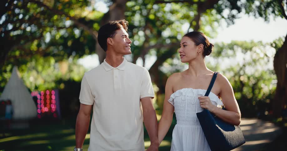 Happy young couple holds hands while taking a romantic walk together through a lush green park