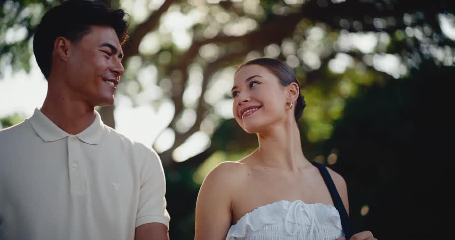 A beautiful young couple smile and gaze at each other lovingly in a park with golden hour sunlight filtering through the trees