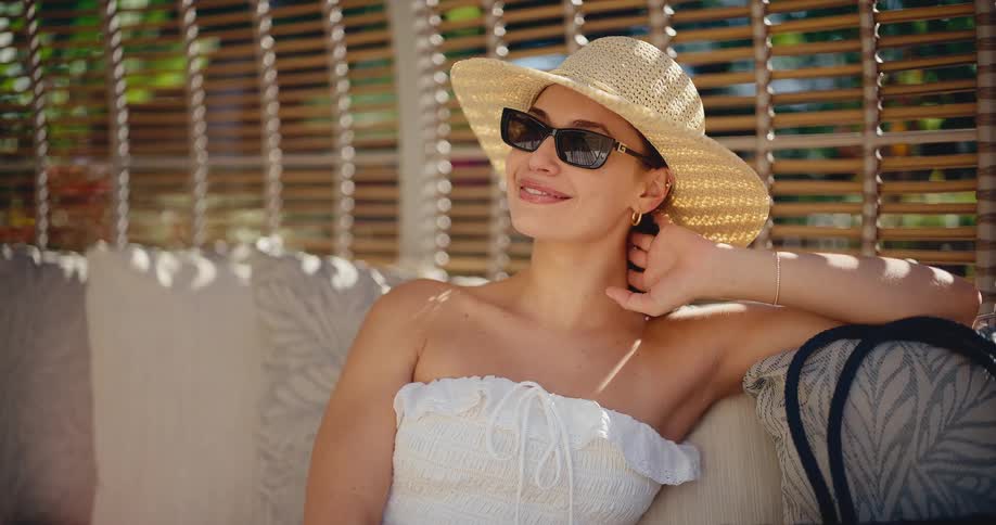An elegant woman in a sun hat and sunglasses smiles while relaxing in a sun-drenched cabana
