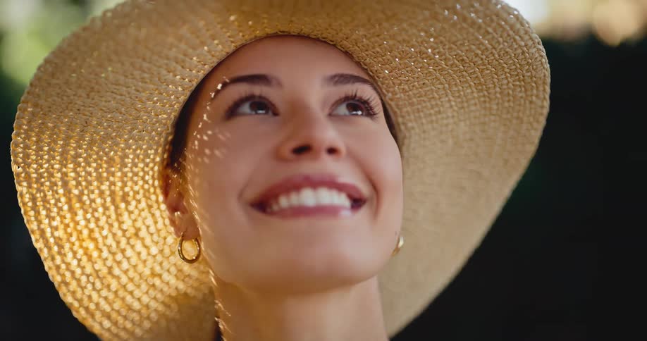 A beautiful woman in a wide-brimmed straw hat smiles serenely while looking up into the warm, dappled sunlight