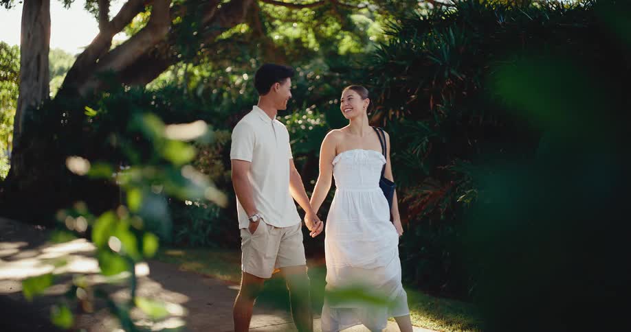 Slow motion view of a happy young couple in white holding hands and walking together down a path in a lush green park