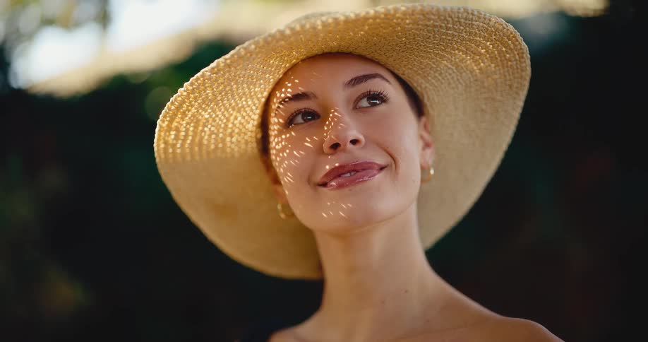 A beautiful young woman in a straw hat smiles contentedly as dappled sunlight dances across her face