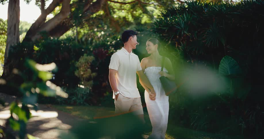 An attractive young couple smiles and holds hands while taking a romantic walk through a lush green park in slow motion