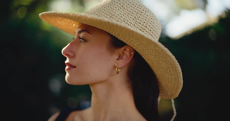 An elegant young woman in a wide-brimmed straw hat gazes thoughtfully while basking in the warm afternoon sunlight