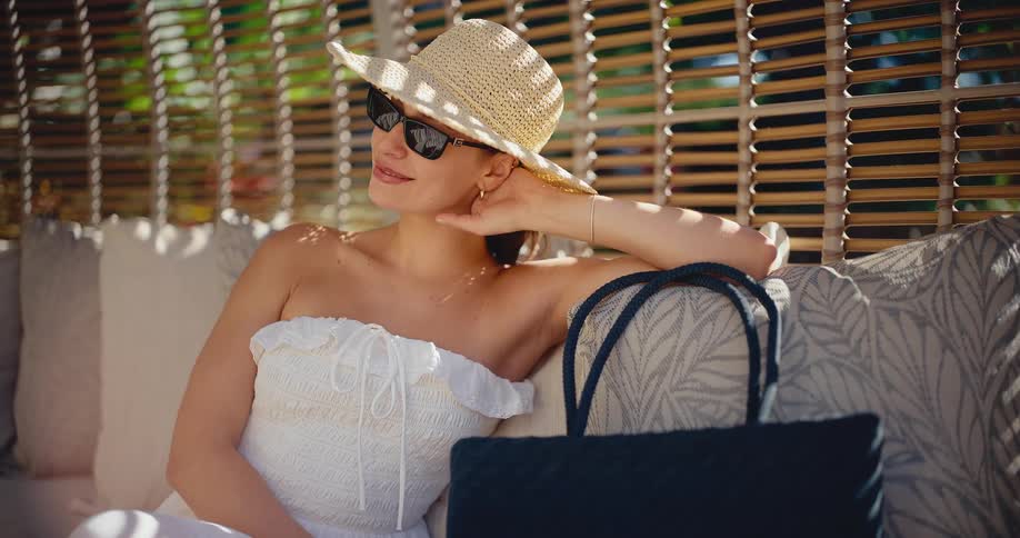 An elegant woman in a white summer dress, straw hat, and sunglasses smiles while relaxing on a luxurious outdoor lounge on a sunny day