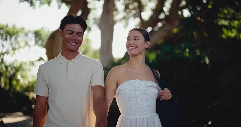 A stylish young couple holds hands and walks together in slow motion through a beautiful tree-lined path during golden hour