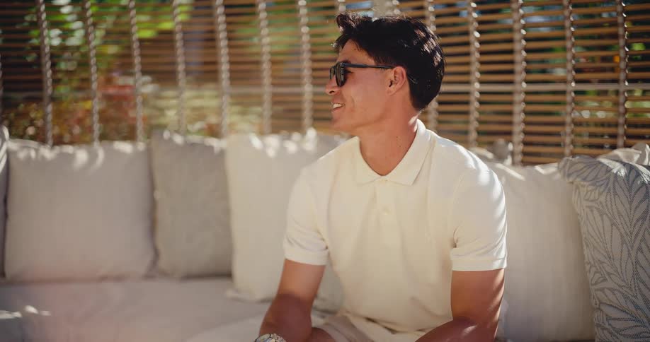 Portrait of a smiling, handsome man in sunglasses turning to the camera while lounging on a sunlit patio couch