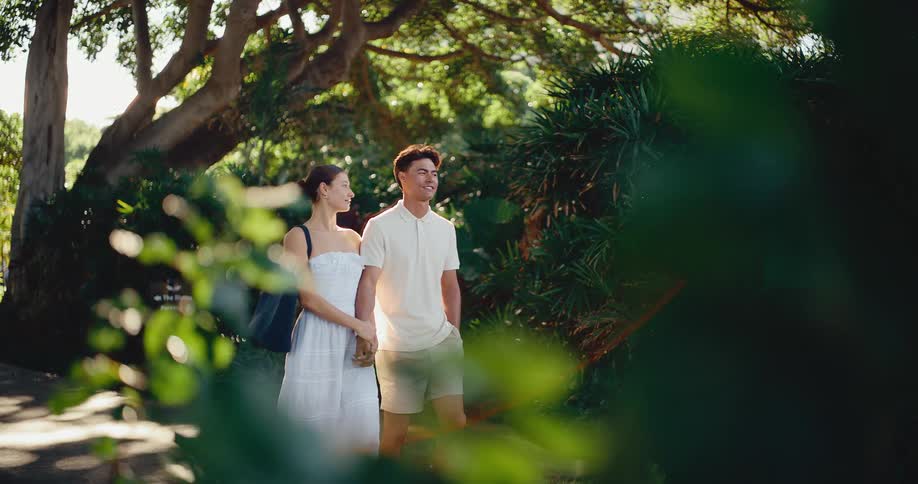 An attractive young couple strolls hand-in-hand through a sun-dappled tropical park on a beautiful day