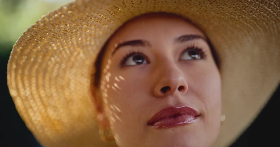 A close-up of a smiling woman in a straw hat looking up as dappled sunlight dances across her face