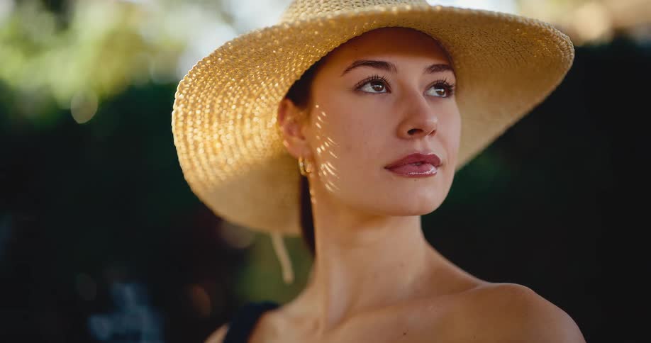 A serene young woman in a wide-brimmed straw hat gazes thoughtfully with dappled sunlight on her face in a lush garden setting