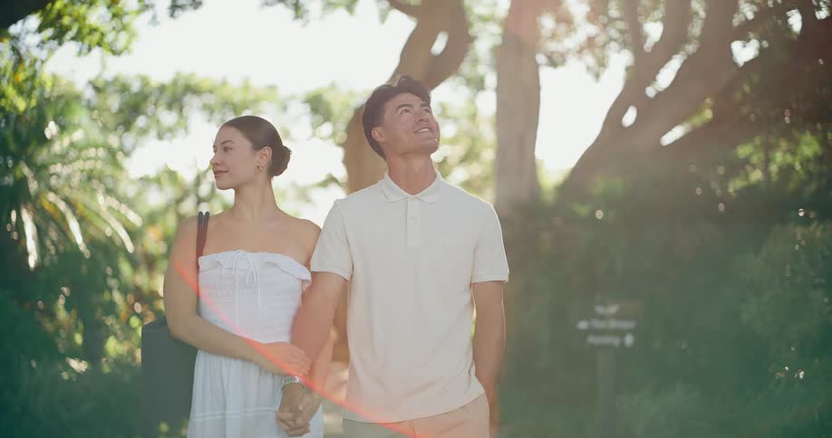 A stylish young couple smiles and holds hands while walking in slow motion along a beautiful tree-lined path at a sunny, tropical resort
