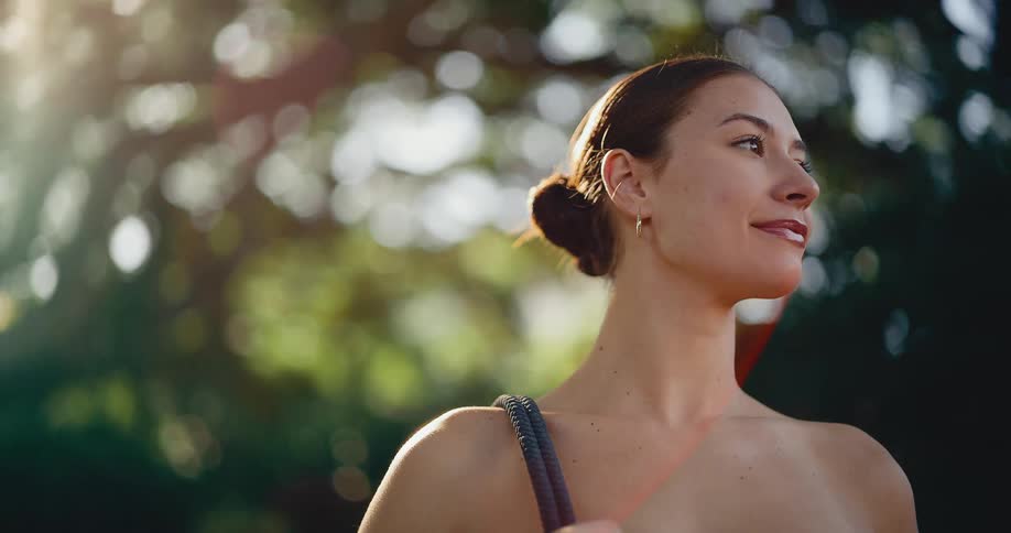 A serene young woman looks up towards the bright sun in a park before turning her head and smiling warmly