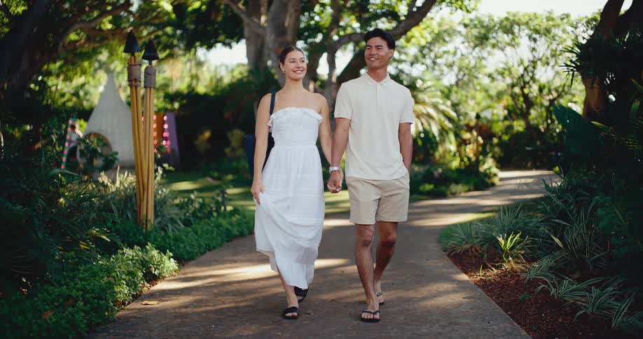 A beautiful couple in love strolls hand in hand down a paved path through a lush, tropical garden on a sunny day