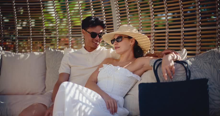 A stylish couple relaxes and chats while lounging in a luxurious cabana on a sunny day at a tropical resort