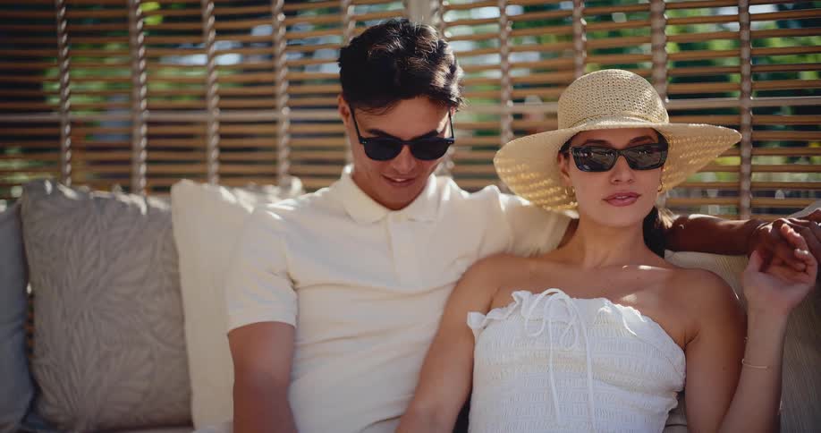 An affectionate couple in white outfits and sunglasses talk and relax together on a sun-drenched resort patio