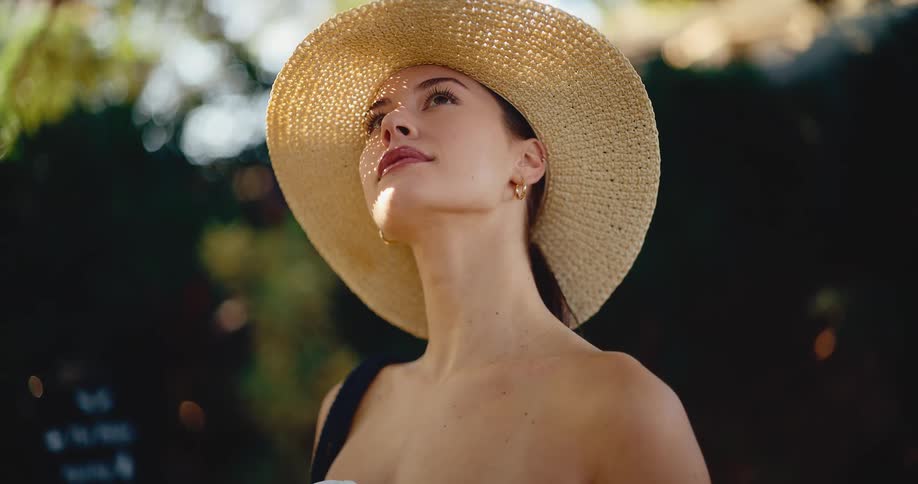 A stylish young woman in a straw sun hat looks up and smiles peacefully as she enjoys the warm, dappled sunlight