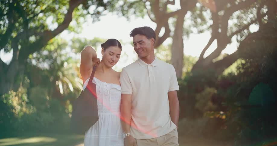 A happy young couple holds hands while taking a romantic walk through a lush, sunlit park in slow motion
