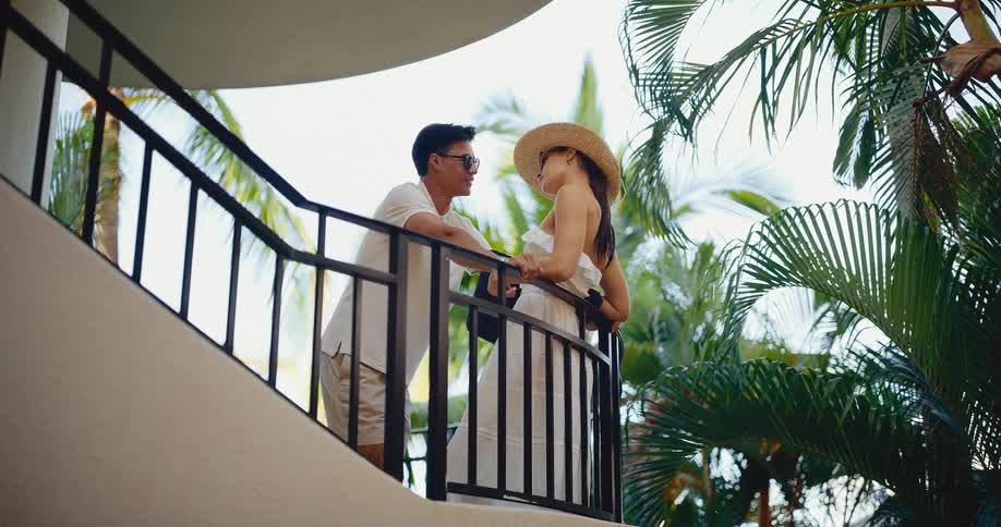 A young, stylish couple talks intimately on a curved hotel balcony surrounded by lush tropical palm trees