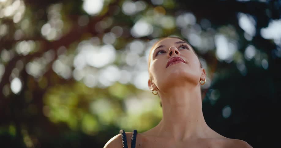 A peaceful young woman looks up contemplatively as golden hour sunlight shines through the lush green trees behind her