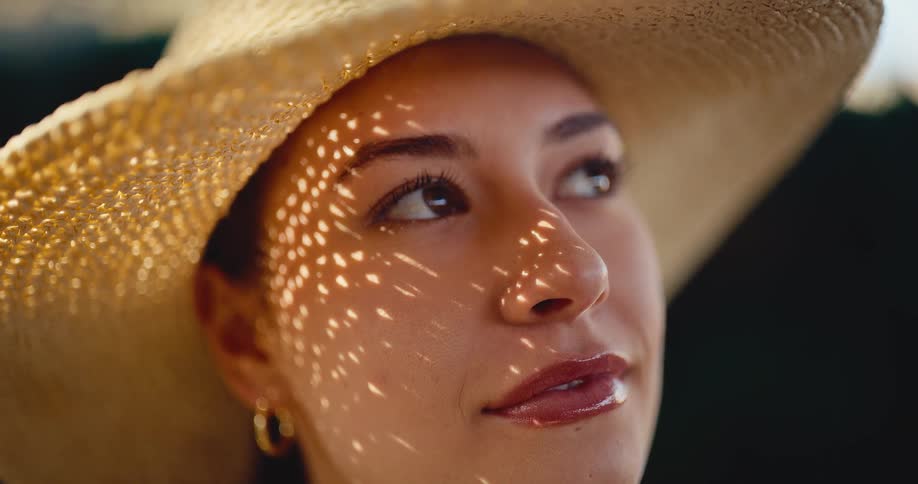 Close-up portrait of a serene woman enjoying the sunshine, with light from her straw hat dappling her smiling face