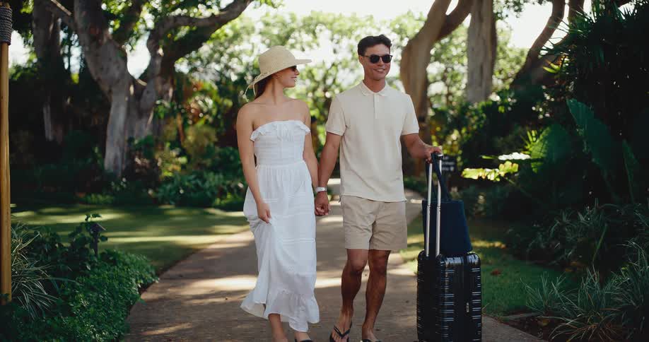 Stylish couple walks hand-in-hand with luggage through a lush tropical resort on vacation