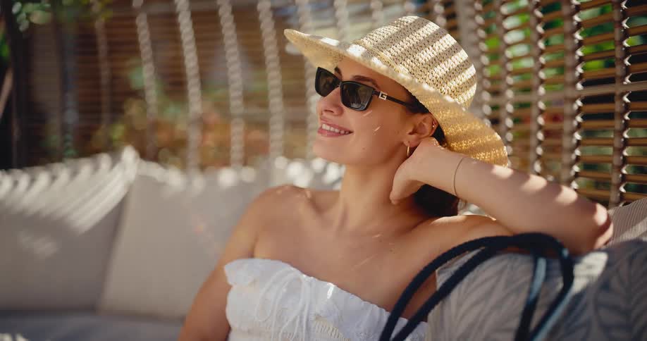 A stylish woman in a straw hat and sunglasses relaxes on a luxurious outdoor lounge, enjoying the warm sunshine