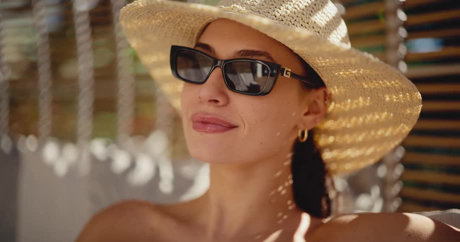 Close up of a smiling woman in a stylish sun hat and sunglasses relaxing in the dappled sunlight on a warm summer day