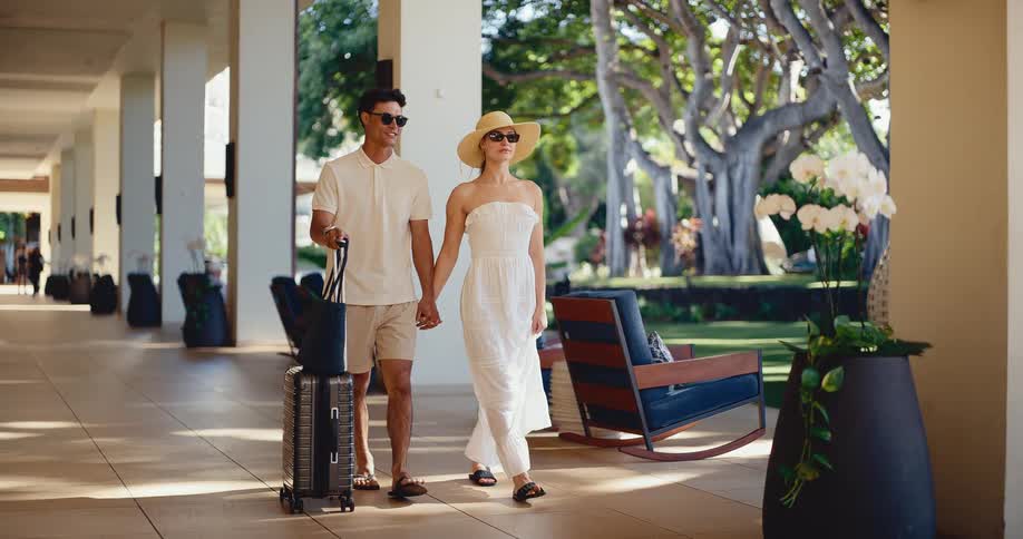 A stylish young couple holds hands while walking with their luggage through the open-air lobby of a luxurious tropical resort