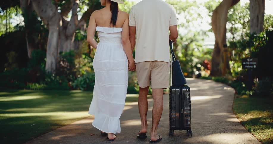 A stylish couple holds hands and walks with their suitcase down a beautiful path, arriving at a luxury tropical resort for their vacation
