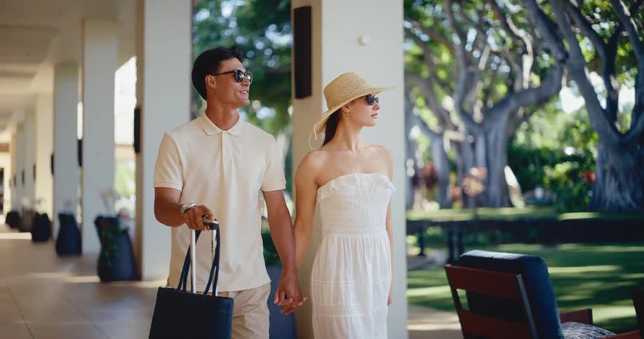 Stylish couple holding hands and walking with luggage arrives at a luxury tropical hotel for their vacation