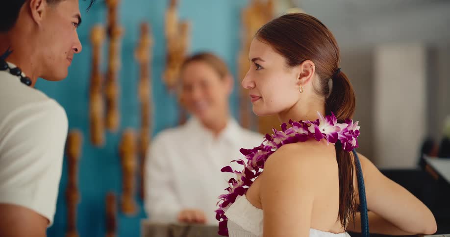 Side view of a beautiful young couple with leis happily checking in at a tropical hotel reception desk