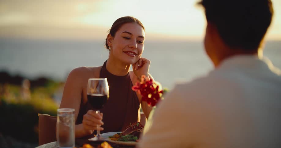 A beautiful woman smiles at her date while enjoying a glass of red wine during a romantic dinner overlooking the ocean at sunset