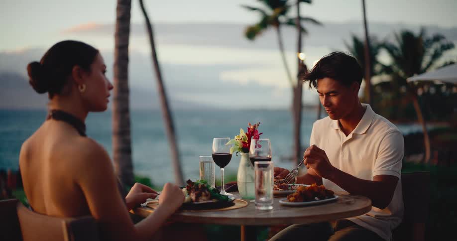 An elegant couple dines together at a beautiful beachfront restaurant as the sun sets over the ocean