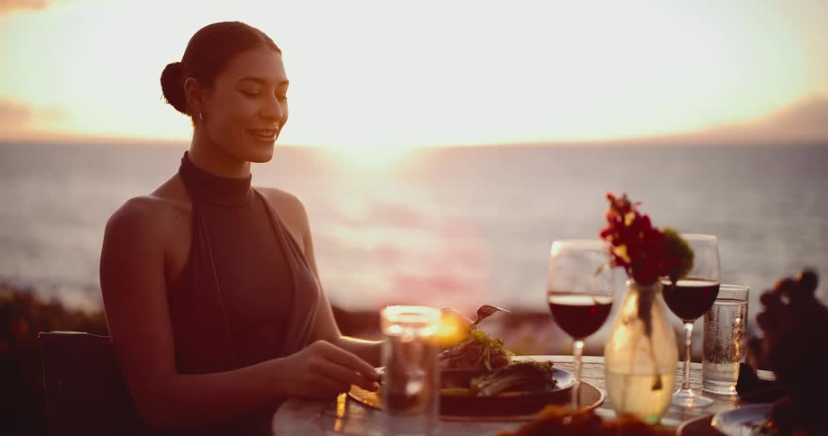 A beautiful young woman enjoys a romantic dinner with wine overlooking the ocean at sunset