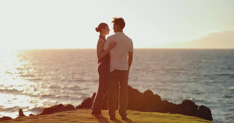 A loving couple embraces on a coastal cliff, watching the golden sunset over the ocean