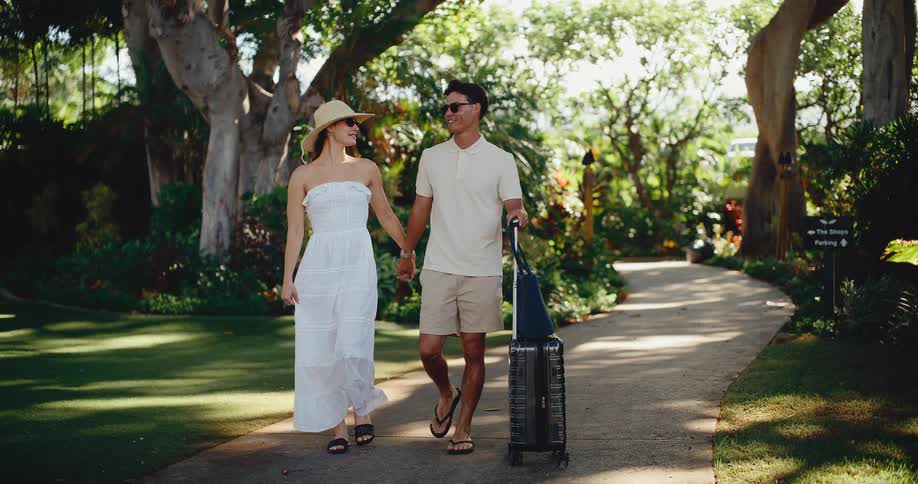 Young attractive couple holding hands and walking with a rolling suitcase through a lush tropical resort, arriving for their vacation