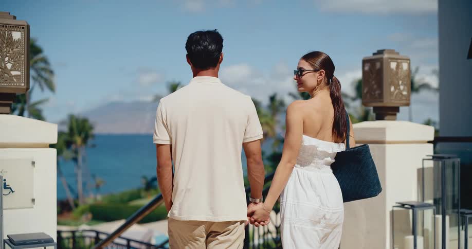 A loving couple holds hands while walking through a beautiful luxury resort with an ocean view on a sunny day