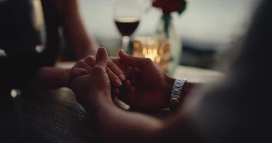 A romantic couple holds hands across a table during an intimate candlelit dinner with wine