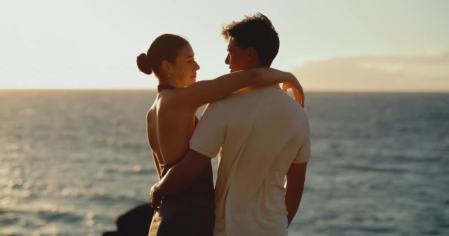 An attractive young couple embrace and look into each other's eyes while watching a beautiful golden sunset over the ocean