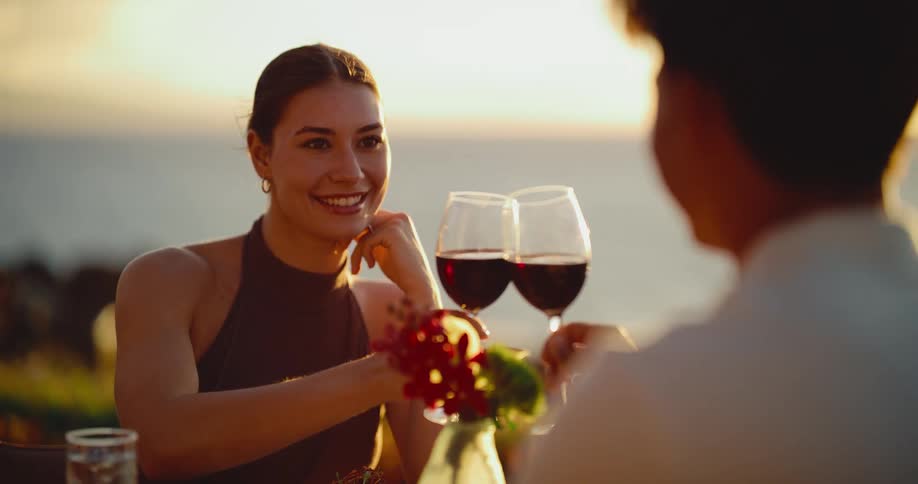 A beautiful young woman smiles and sips red wine on a romantic dinner date with her partner overlooking the ocean at sunset