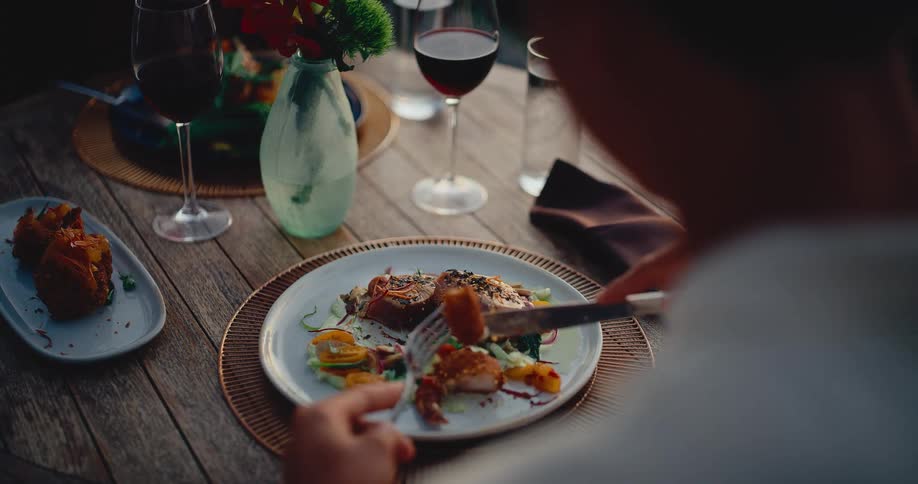 A person savors a gourmet meal with red wine at a rustic outdoor table in slow motion