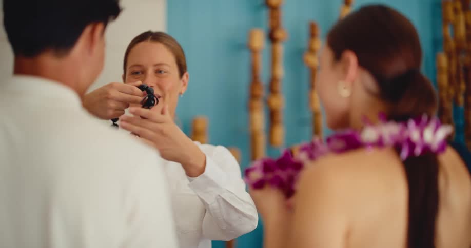 A friendly hotel concierge welcomes a young couple by placing a traditional purple flower lei on the woman and a black kukui nut lei on the man