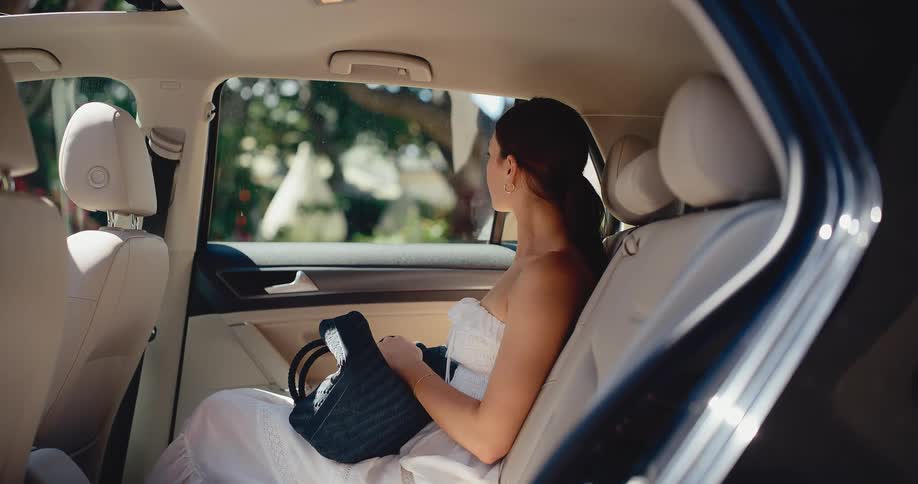 A beautiful woman in a white summer dress sits peacefully in the back of a car on a sunny day