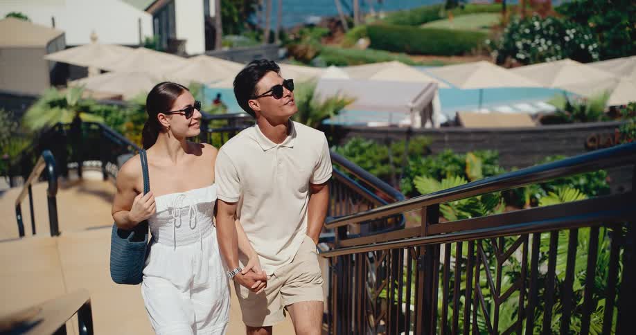 An attractive, stylish couple walks hand-in-hand up a ramp at a beautiful tropical resort in slow motion