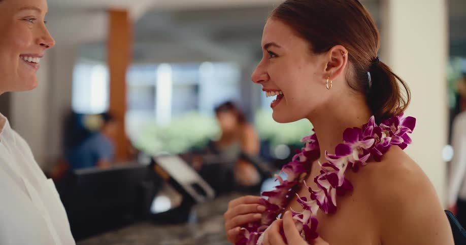 A friendly hotel employee welcomes a smiling young guest by placing a beautiful purple orchid lei around her neck in a tropical resort lobby