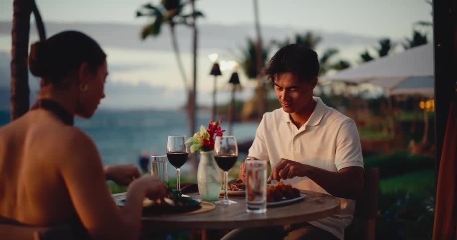A romantic couple enjoys an intimate dinner with red wine at an outdoor table overlooking the ocean at sunset