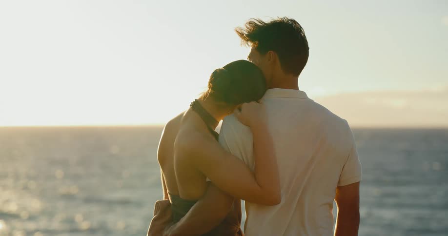A young couple in love embraces while watching a beautiful golden sunset over the ocean