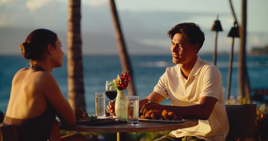 A smiling young couple enjoys a romantic dinner with wine at an oceanfront restaurant during a beautiful sunset
